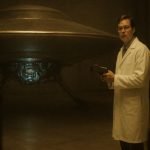 Bob Lazar con bata de laboratorio dentro de un hangar en el Área S-4, examinando el sistema de propulsión de un platillo volante.