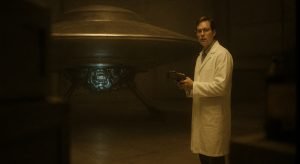 Bob Lazar con bata de laboratorio dentro de un hangar en el Área S-4, examinando el sistema de propulsión de un platillo volante.