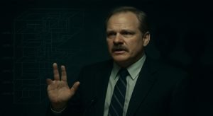 Phil Schneider dando una conferencia, mostrando su mano mutilada, con una superposición de un diagrama de la Base de Dulce y siluetas de Grises, representando su testimonio.